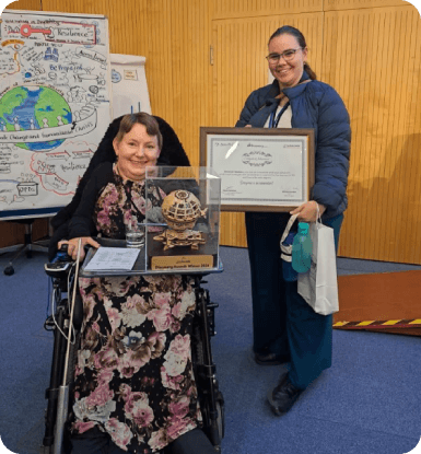 Two women smiling and posing with a framed certificate and a trophy at an award event, with a colorful illustrated board and wooden panel wall in the background.