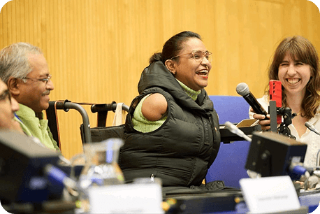 A woman smiling and speaking into a microphone during a panel discussion, with two other participants seated beside her in a professional conference setting.