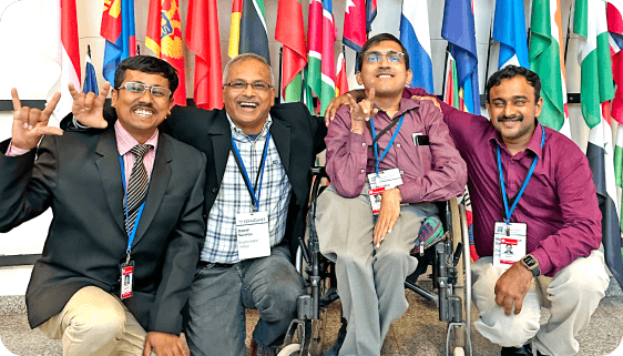 Four smiling men, including one in a wheelchair, posing together in front of international flags during the Discovery Awards event, symbolizing unity, diversity, and achievement.